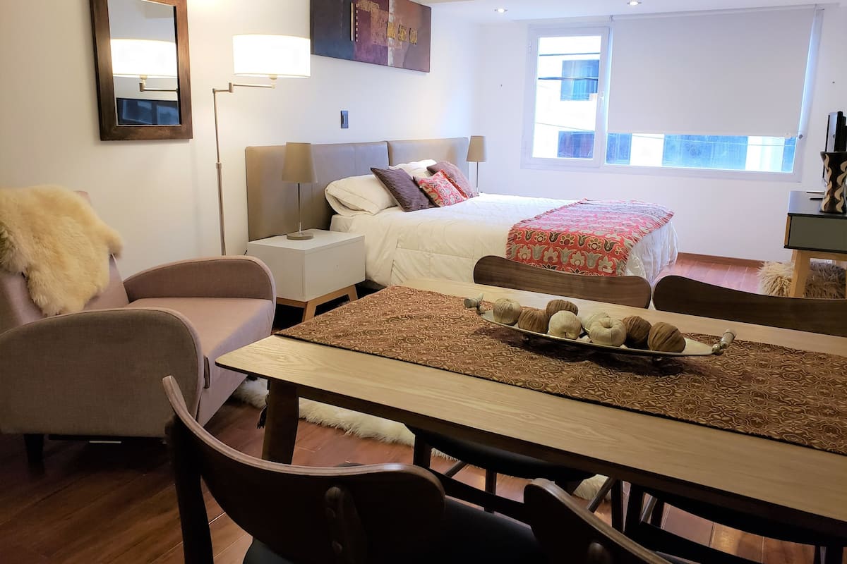 Hotellihuone, jossa on sänky, yöpöytä, lamppu ja ruokapöytä tuoleineen.