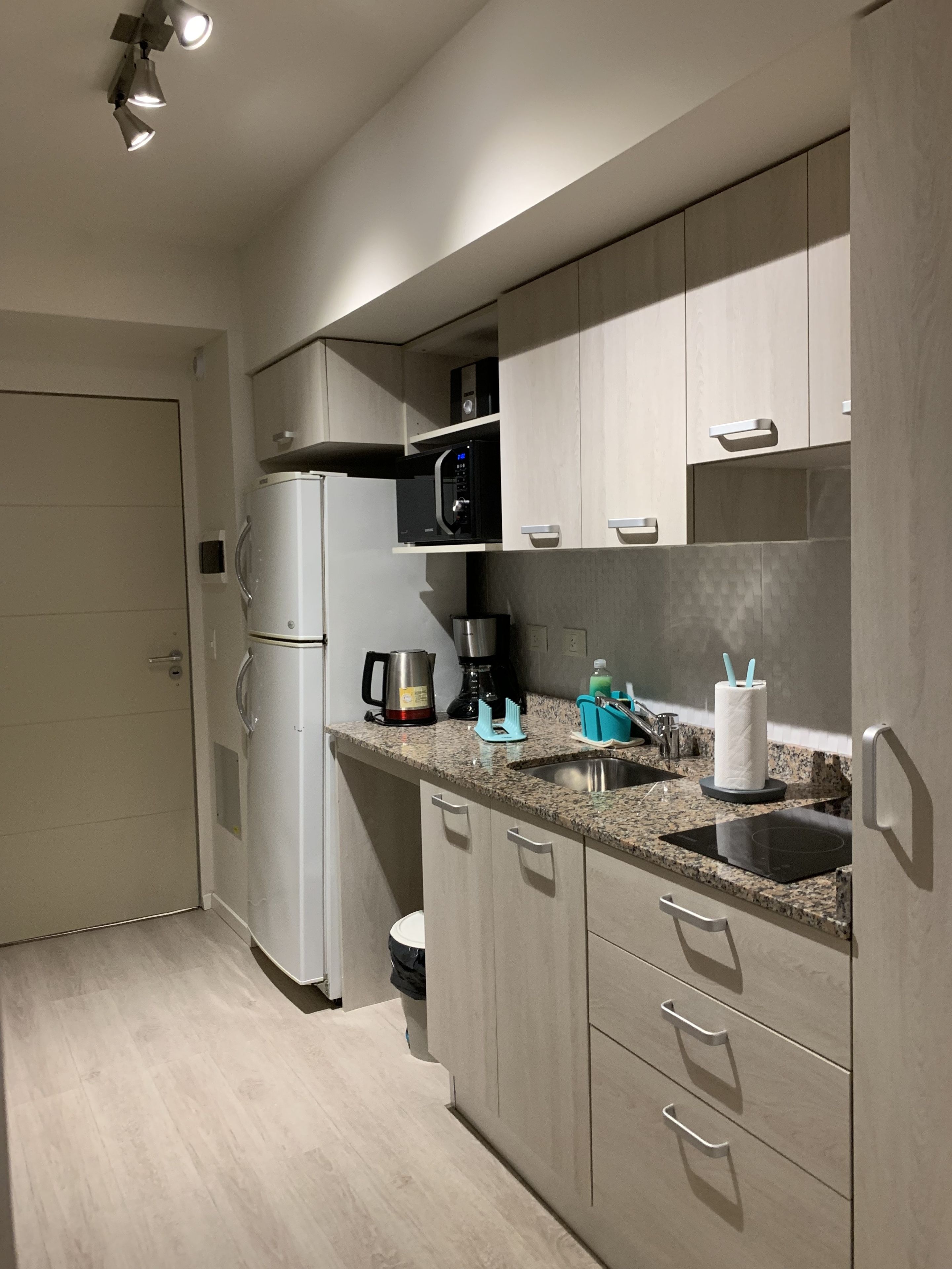 A modern kitchen with a built-in microwave, coffee maker, and kettle. There is a sink with a faucet, a trash bin, and a countertop with a paper towel holder and utensils.
