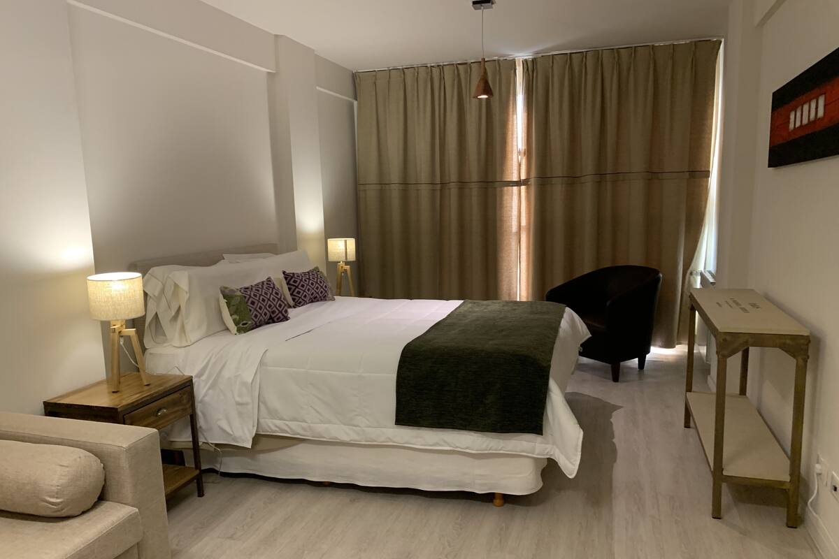Hotellihuone, jossa on sänky, yöpöytälamput, tuoli, pieni pöytä ja ikkuna, jossa on verhot.