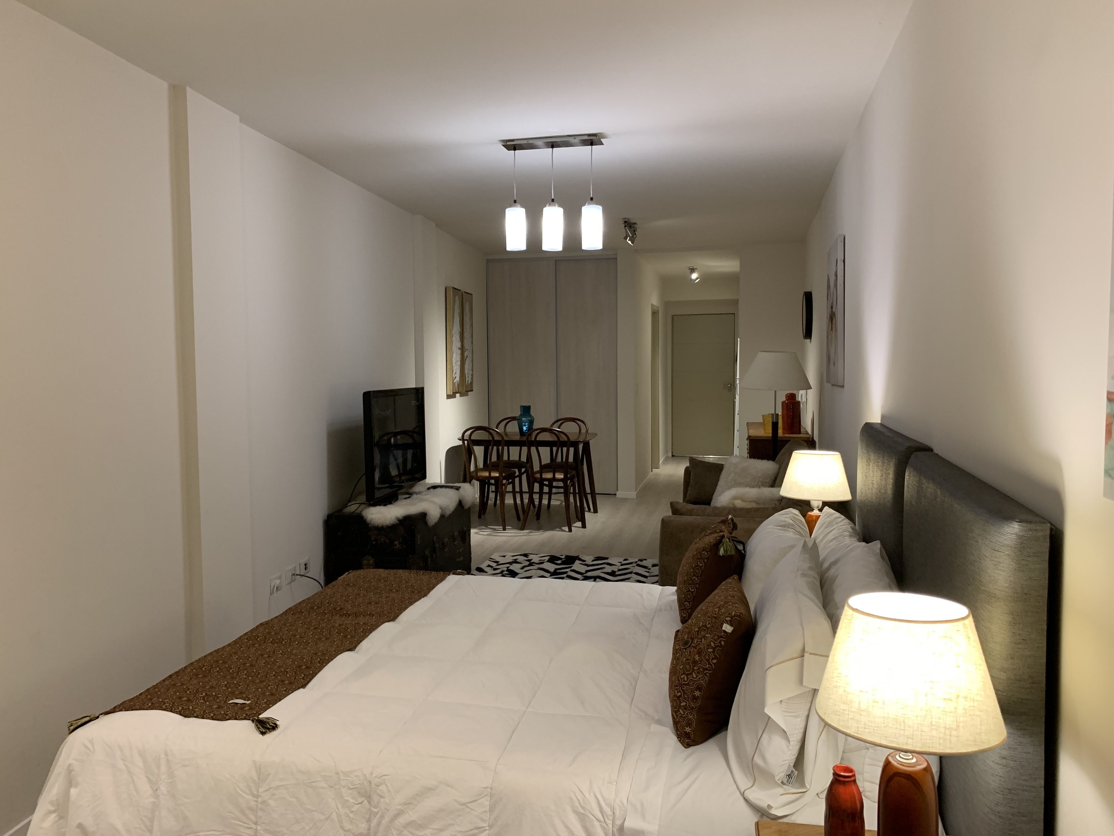Habitación de hotel con cama, lámparas de noche, televisor, escritorio con silla y zona de comedor con sillas y mesa.