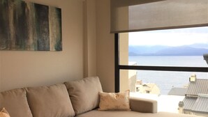 A modern living room with a large sectional sofa, a painting on the wall, and a view of the sea through a large window. - Urbana Suites & Studios 440 (San Carlos de Bariloche)