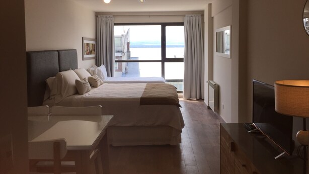 A hotel room with a bed, a desk, a TV, and a view of the sea. - Urbana Suites & Studios 440 (San Carlos de Bariloche)