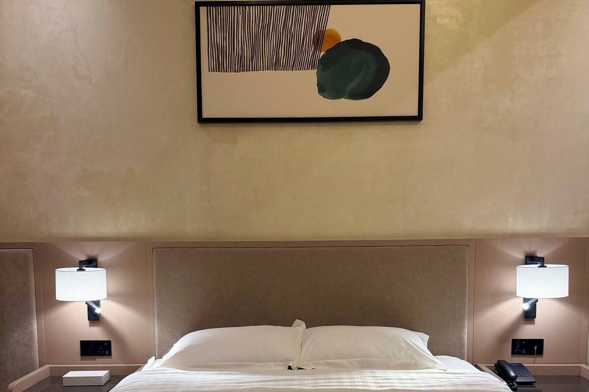A hotel room with a large bed, two bedside tables with lamps, a control panel, and a framed abstract artwork on the wall.