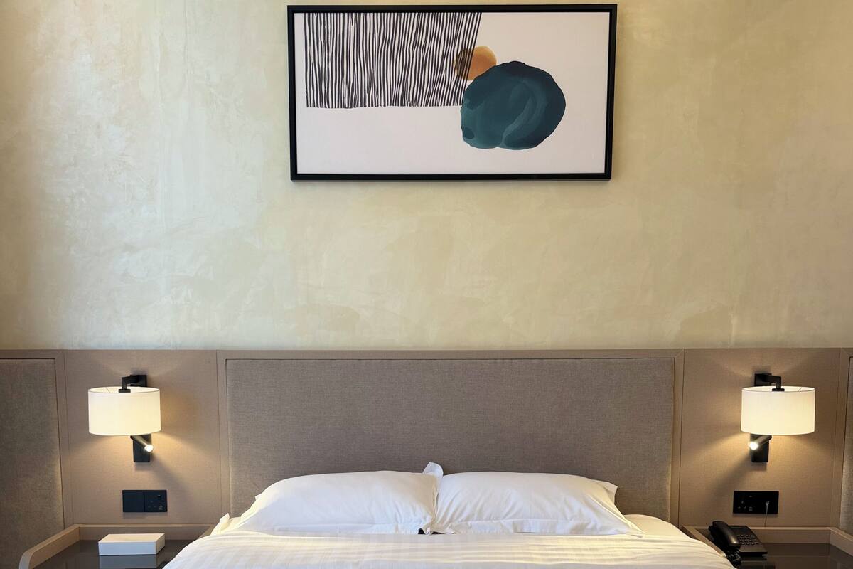 A hotel room with a large bed, two bedside lamps, a framed abstract artwork above the bed, and a nightstand with a phone and a box.