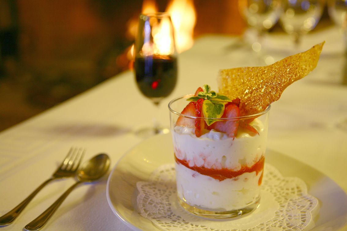 Un dessert servi dans un verre, composé de fraises et d’une gaufrette, présenté sur une assiette blanche ornée d’une serviette en dentelle, accompagné d’une fourchette et d’une cuillère, et agrémenté d’une bougie en arrière-plan.