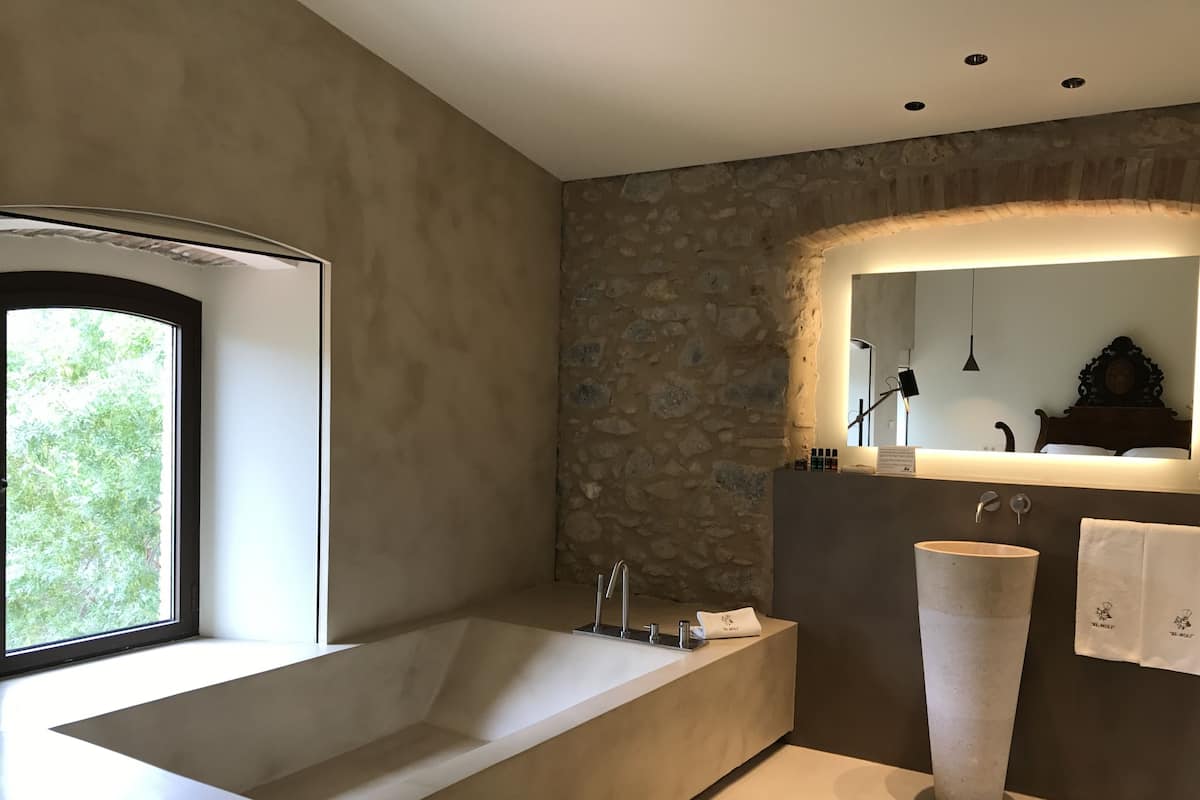 Un baño moderno con una bañera grande, una pared decorativa de piedra, un espejo amplio y un lavamanos alto y cilíndrico.