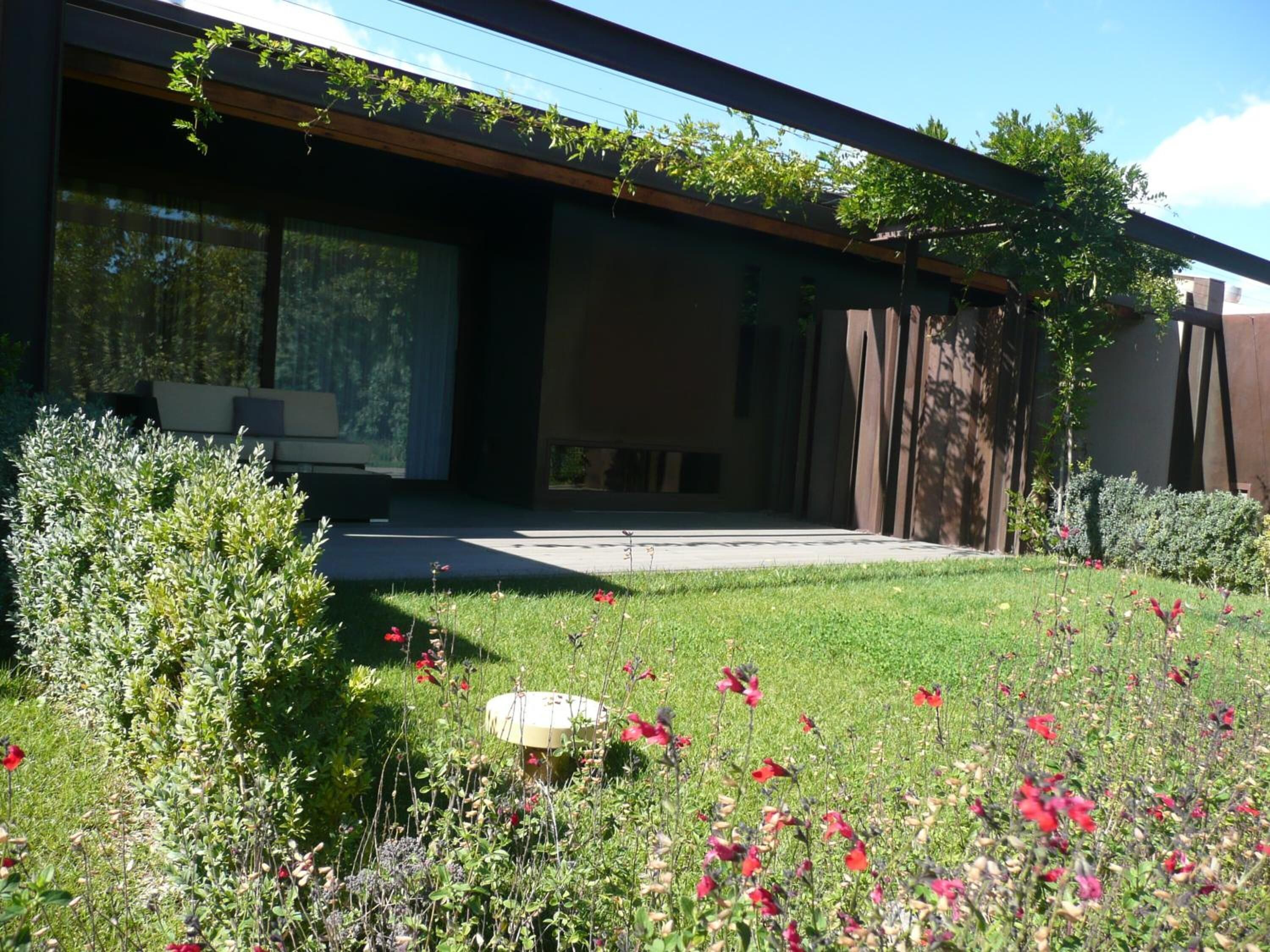 Un edificio moderno con grandes ventanales, un patio cubierto y un jardín bien cuidado con diversas plantas y flores.