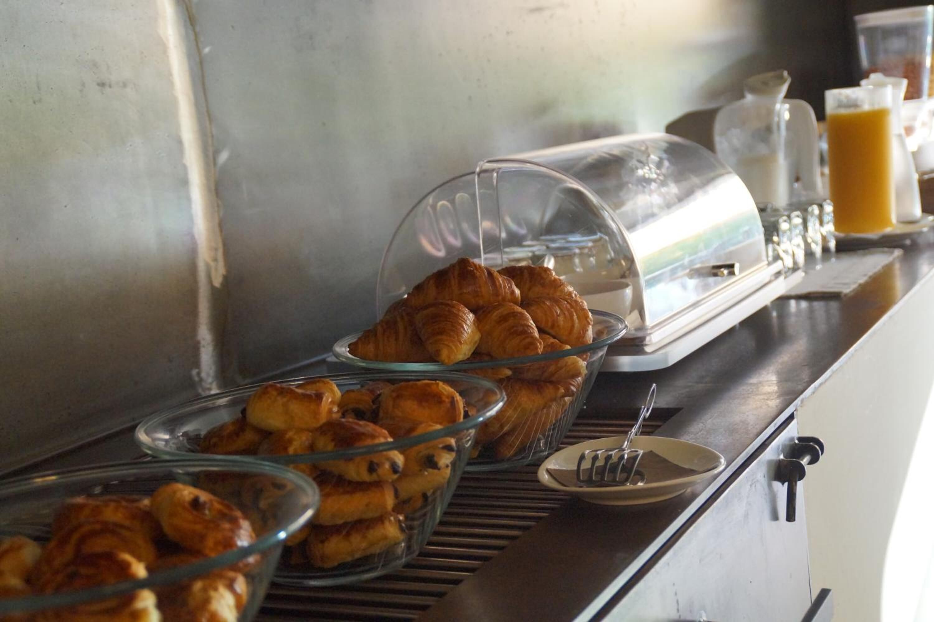 A breakfast buffet with croissants and pastries in glass bowls, a coffee machine, and a glass of orange juice.