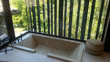 Une salle de bain moderne avec une baignoire sur pieds, un petit tabouret et une vue sur un jardin verdoyant, visible à travers une fenêtre dotée de stores verticaux.