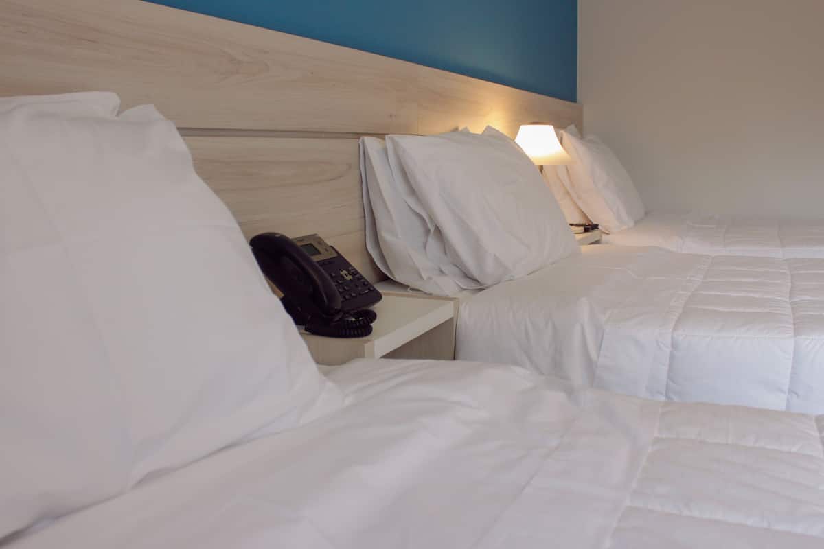 Habitación de hotel con dos camas, ropa de cama blanca, teléfono junto a la cama y mesita de noche con lámpara.