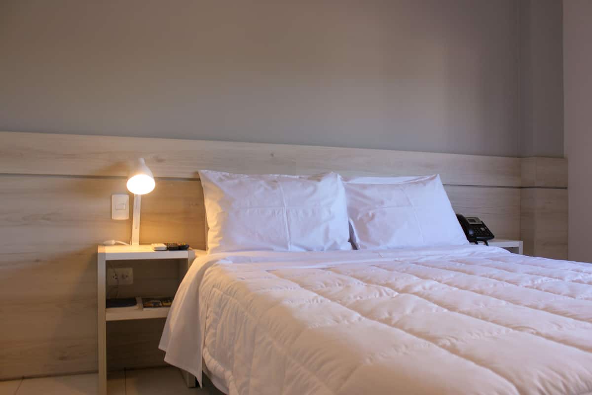 Una cama bien hecha con ropa blanca y almohadas, una lámpara de noche y un teléfono en la mesita.