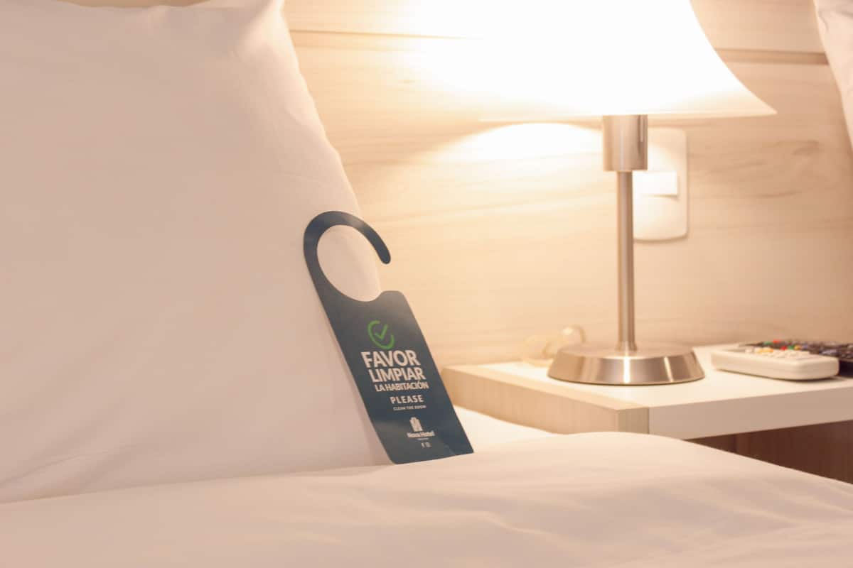 Habitación de hotel con cama, mesita de noche, lámpara y un cartel que invita a los huéspedes a ordenar.
