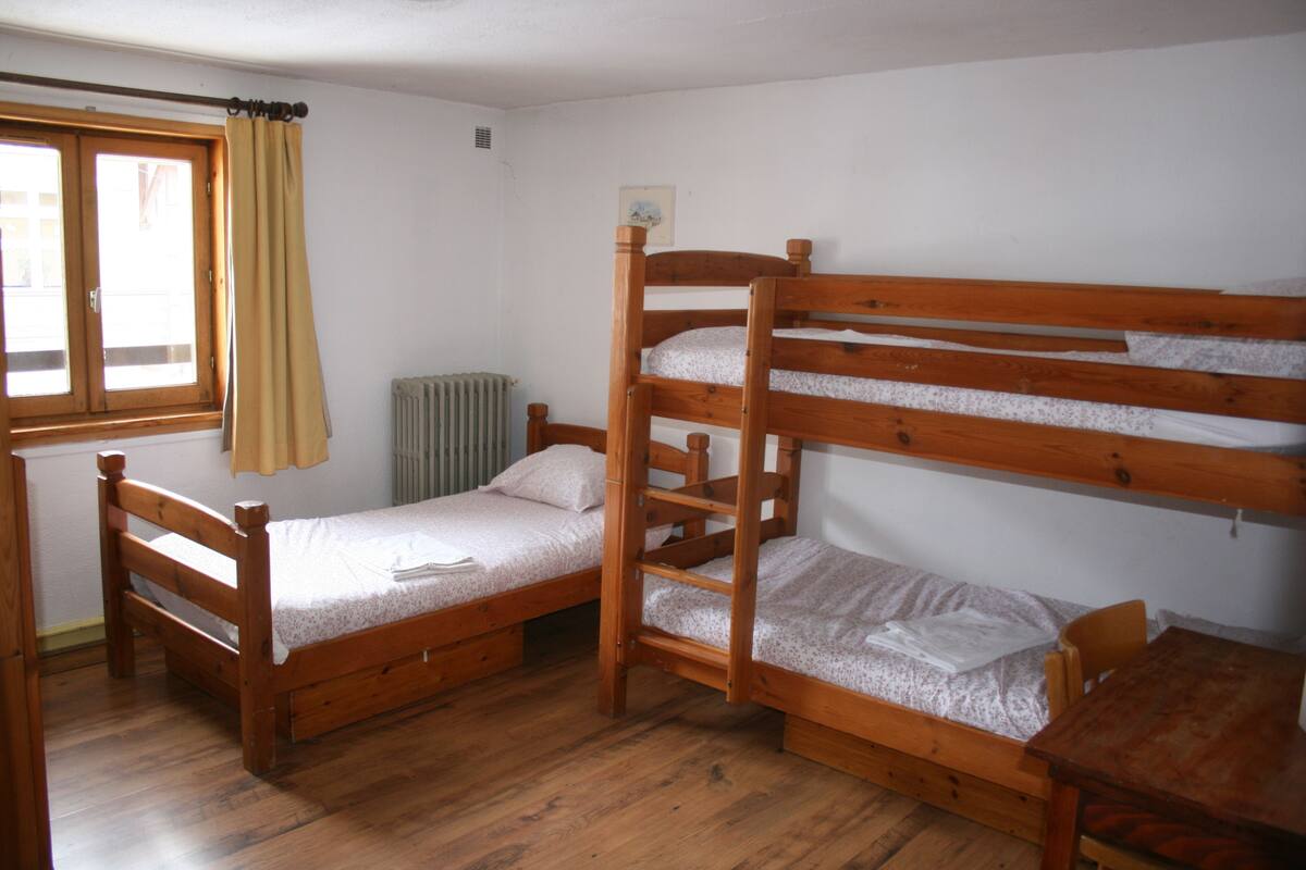 Une pièce avec deux lits superposés en bois, une fenêtre avec des rideaux, un radiateur et une table en bois.
