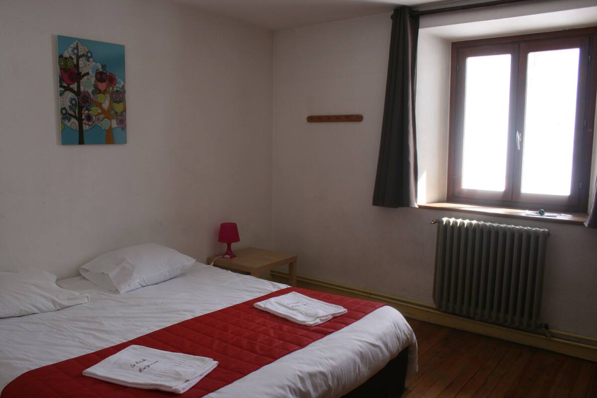 Une chambre d’hôtel avec un lit, une fenêtre avec des rideaux, un radiateur, une petite table avec une lampe et un tableau au mur.