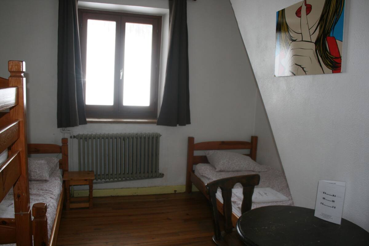 Une chambre de dortoir avec deux lits superposés en bois, une petite table, un radiateur et une fenêtre avec des rideaux.