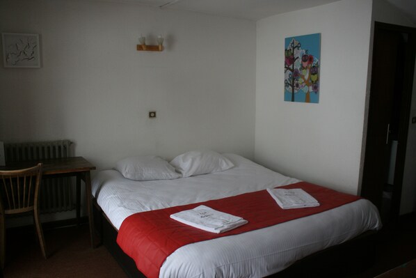 A single bed with a red blanket, a wooden chair, a small table, and a wall with a painting.