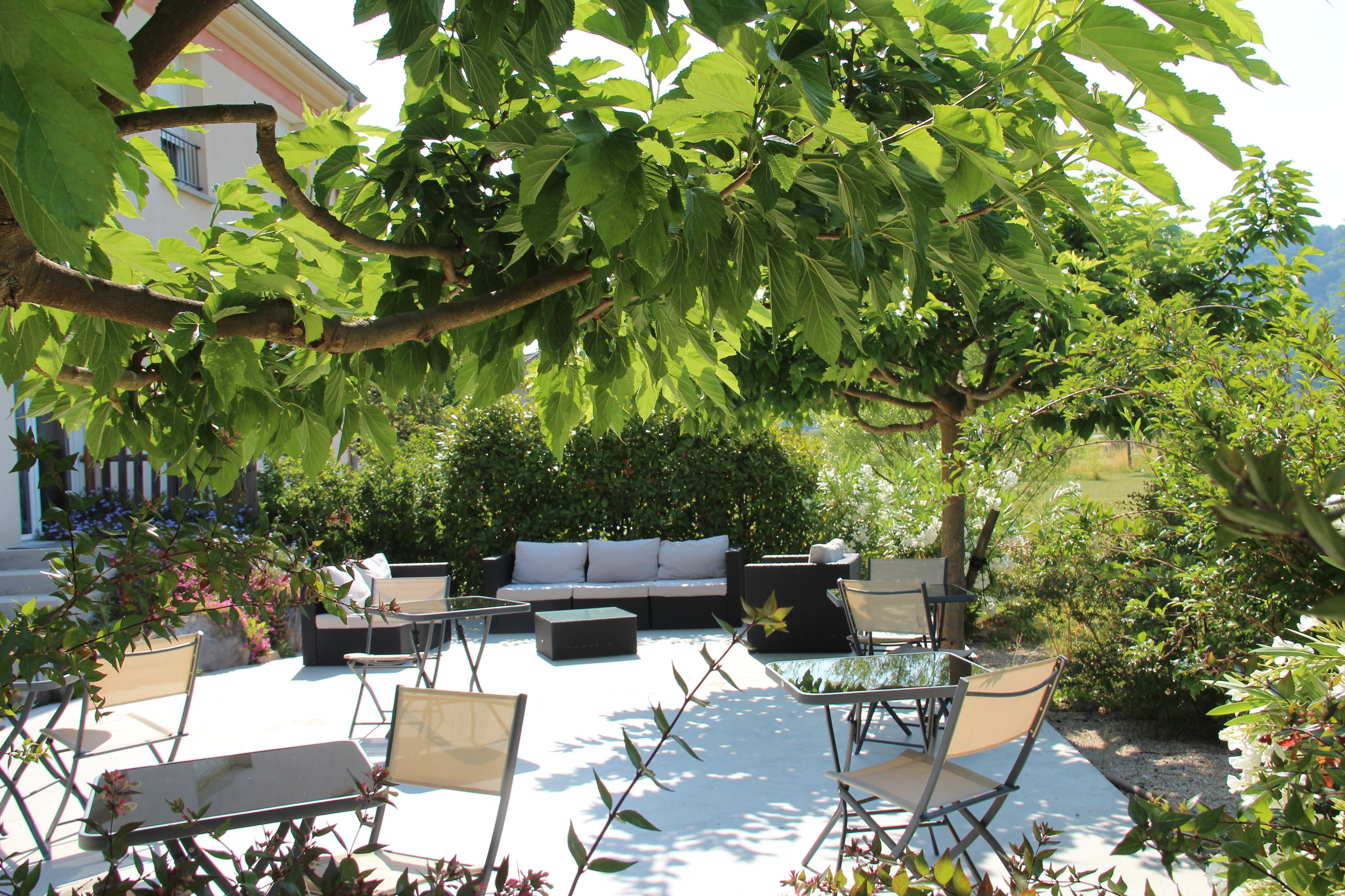 An outdoor seating area with a sofa, chairs, and a table, surrounded by greenery and a building in the background.