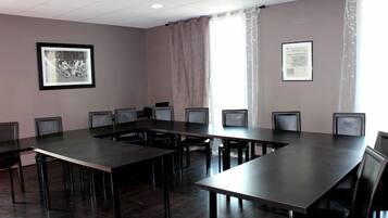 Un salón de conferencias con una gran mesa rectangular rodeada de sillas, un cuadro en la pared y una ventana con cortinas.