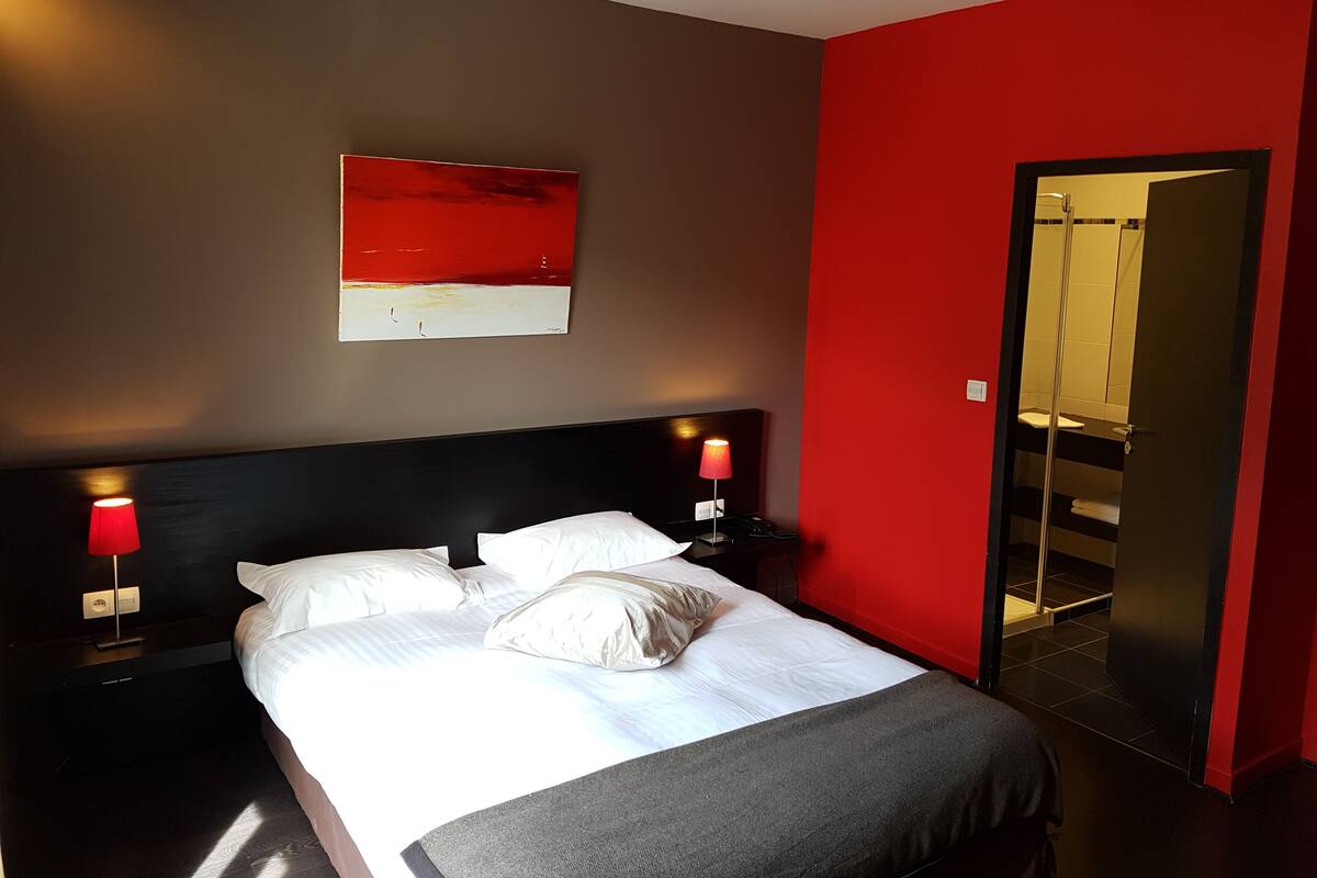 A hotel room with a bed, two bedside lamps, a red accent wall, and a large mirror.