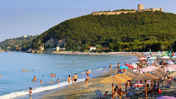 A beach with umbrellas and people, a castle on a hill, and a clear sky.