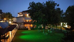 A two-story building with a flat roof and a balcony. There is a well-maintained lawn with a green-lit area and outdoor furniture.