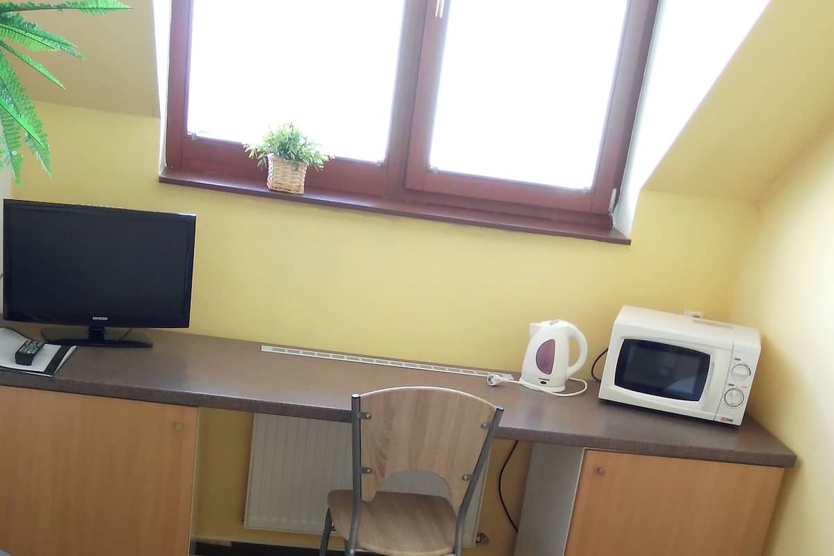 Une petite pièce comprenant un bureau, une chaise, un lit, un téléviseur, un four à micro-ondes et une fenêtre agrémentée d’une plante.