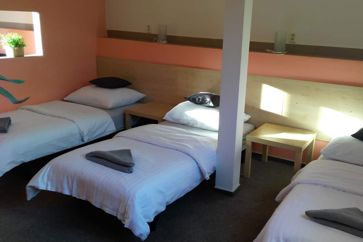 Une chambre d’hôtel avec trois lits simples, chacun muni d’un oreiller et d’une serviette pliée. Une table de chevet en bois avec une lampe se trouve entre les lits. La chambre est dotée d’une fenêtre donnant sur l’extérieur.