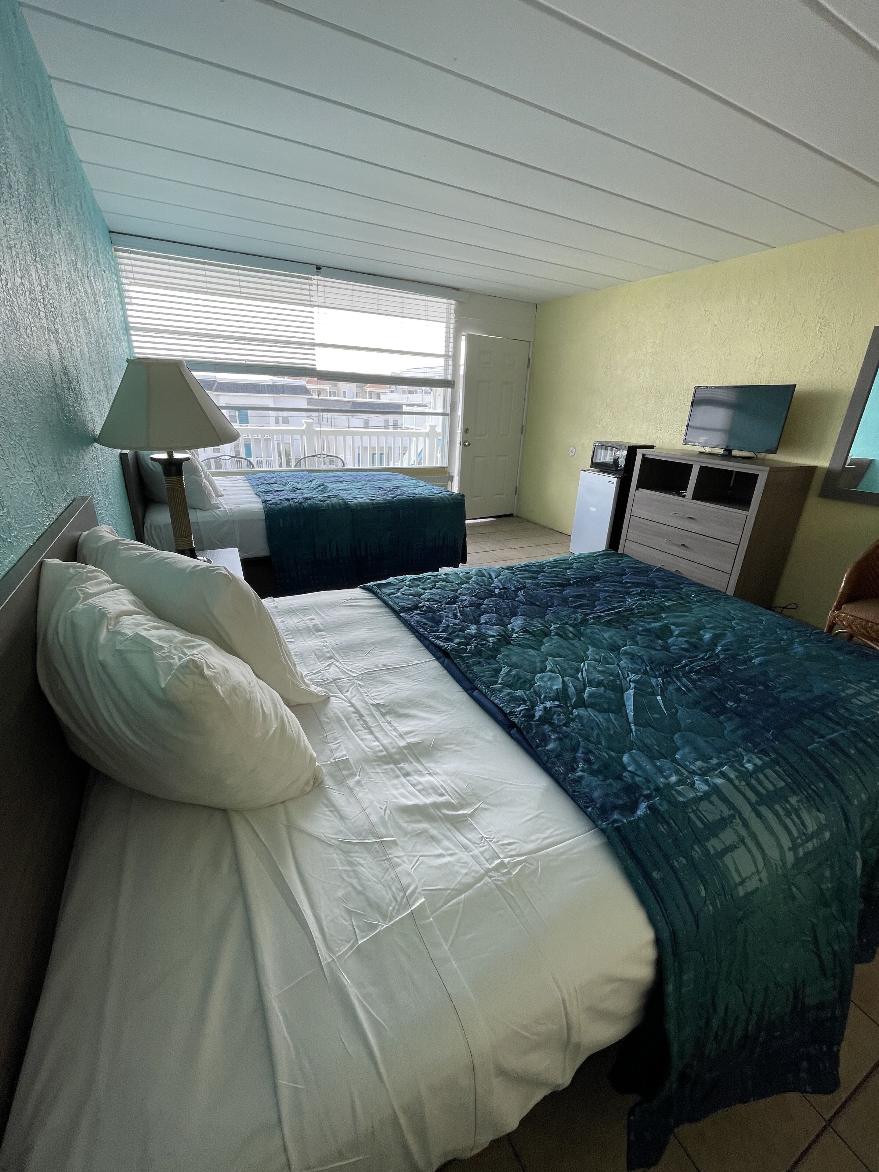 A hotel room with two beds, a TV, a desk, and a window with blinds.