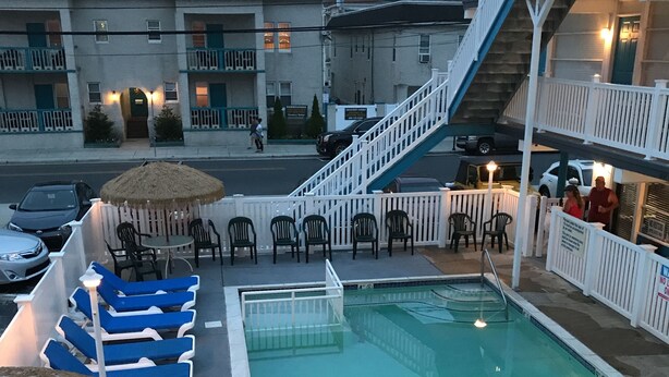 A pool area with lounge chairs and a thatched umbrella, surrounded by a white fence. - Bird Of Paradise Motel (North Wildwood)