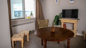 Ein Wohnzimmer mit einem Holzesstisch, einem Fernseher auf einem Holzständer, einem Stuhl, einem kleinen Tisch und einem Fenster mit Meerblick.