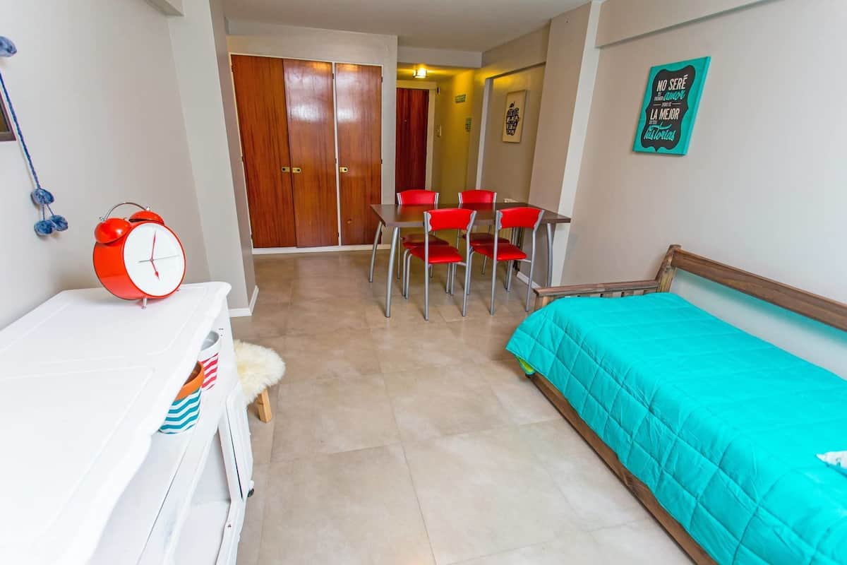 A room with a bed, a table with chairs, a red alarm clock, and a door in the background.
