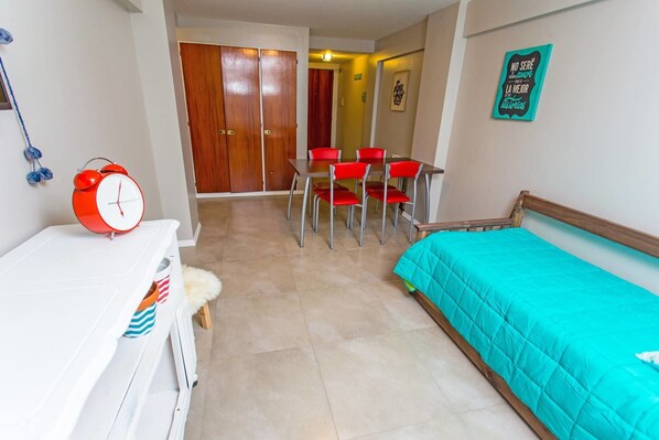 Um quarto com uma cama, uma mesa com cadeiras, um despertador vermelho e uma porta ao fundo.