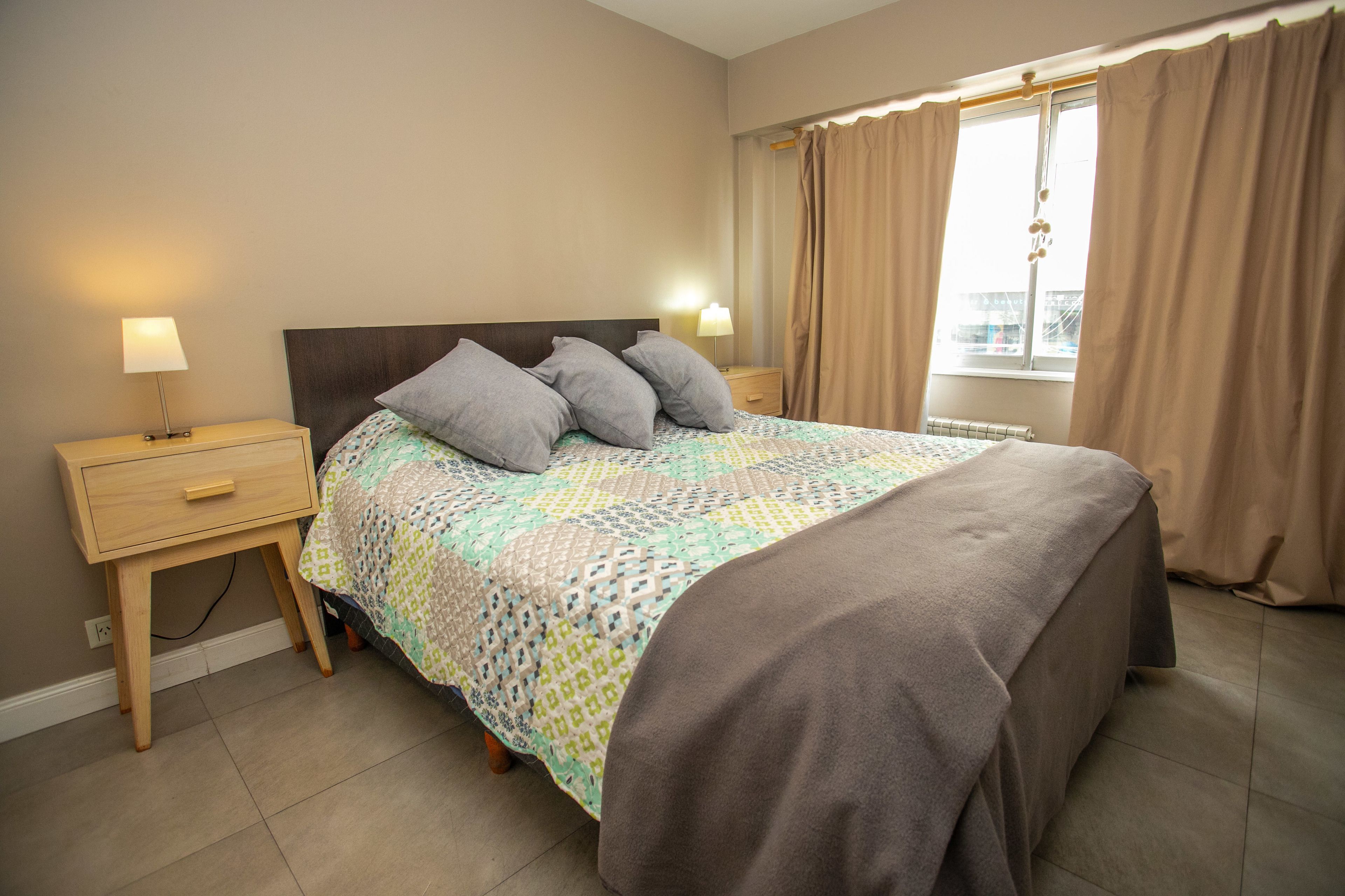 Une chambre à coucher avec un lit, deux tables de chevet avec des lampes, une fenêtre avec des rideaux et un radiateur.