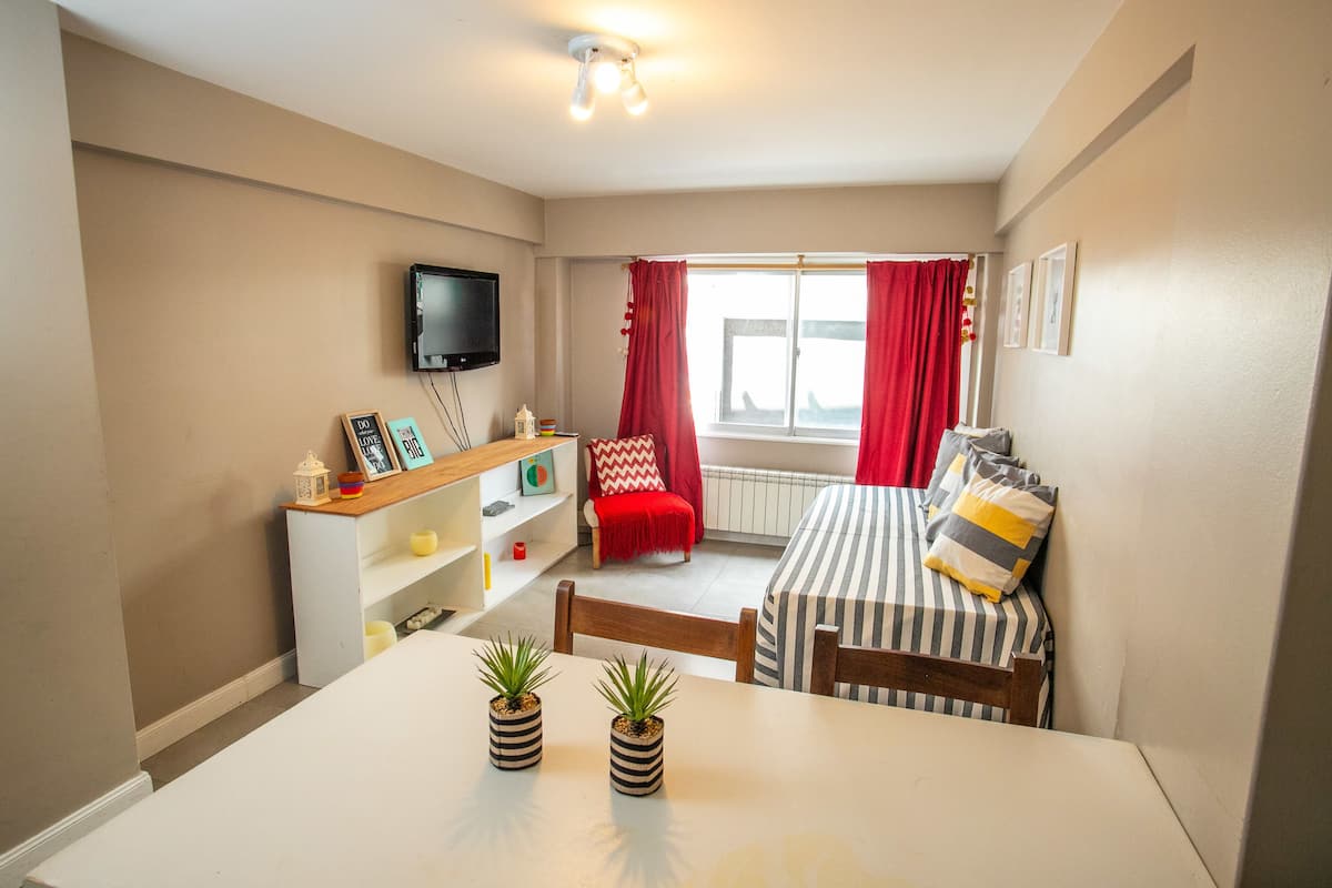 A compact living space with a dining table, two chairs, a TV mounted on the wall, a window with red curtains, and a small bookshelf.