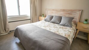 Ein Schlafzimmer mit einem Bett, einem Nachttisch, einem Fenster mit Vorhängen und einer kleinen Pflanze auf dem Nachttisch.