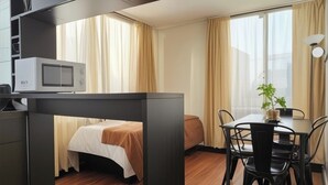 Ett kompakt hotellrum med ett litet kök, en säng, ett matbord med stolar och en krukväxt.