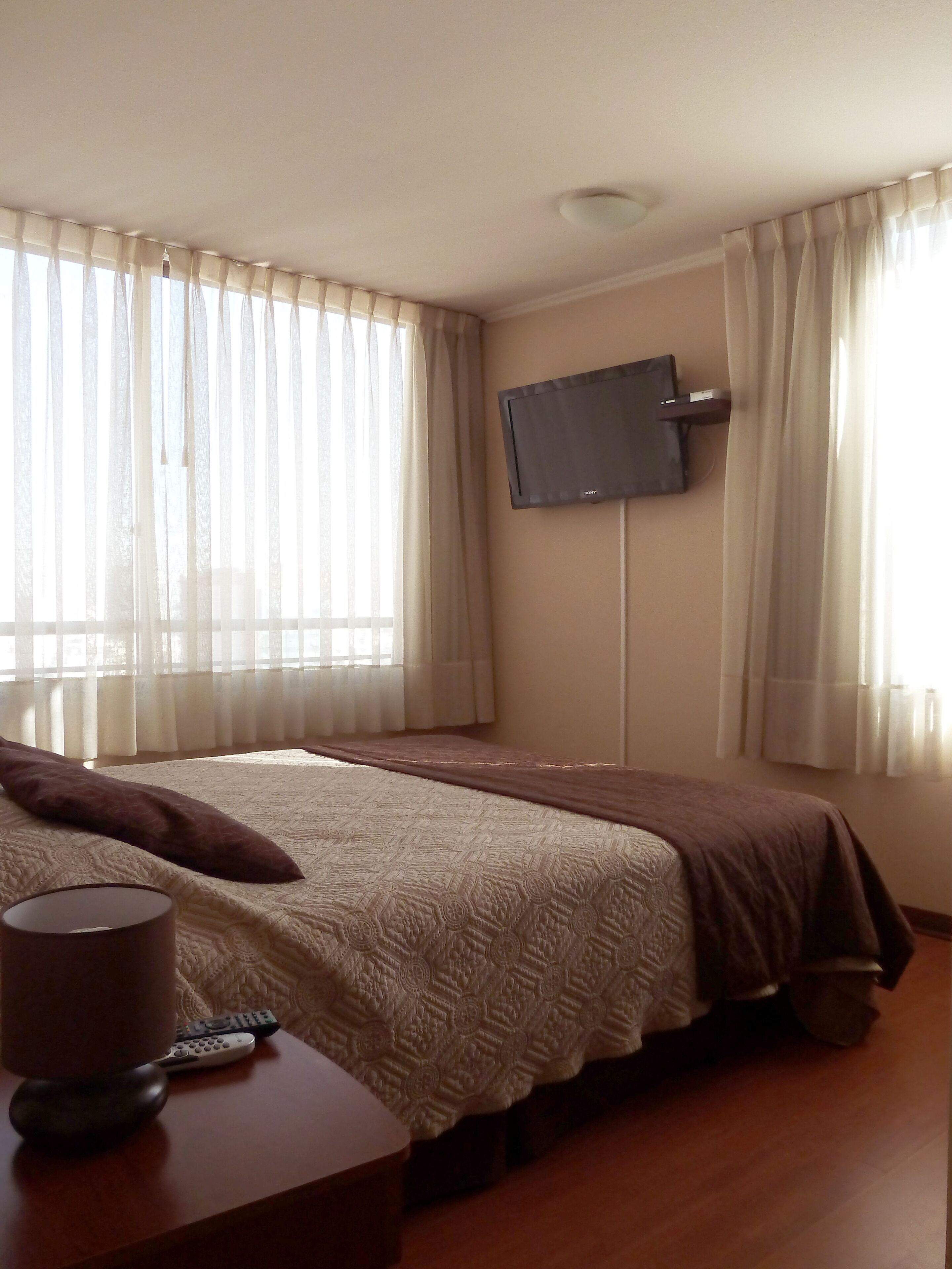 Habitación de hotel con cama, televisor montado en la pared y ventana con cortinas.
