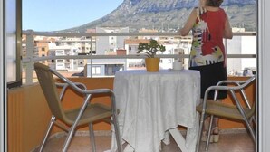 A balcony with a table, chairs, and a potted plant. A person is standing by the window, overlooking a mountainous landscape. - Hostal Comercio (Denia)