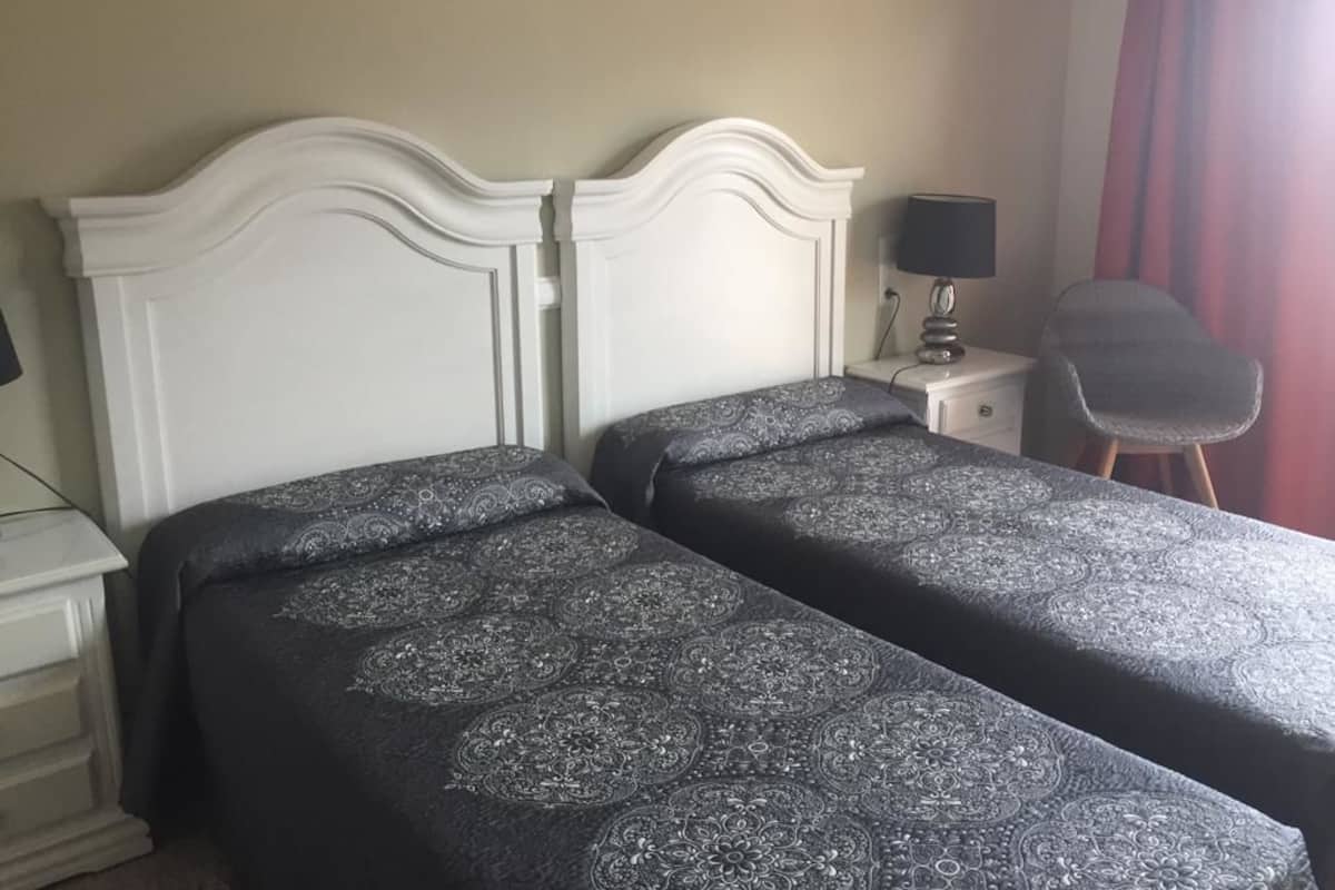 A twin room with two beds, a bedside table with a lamp, a chair, and a window with curtains.