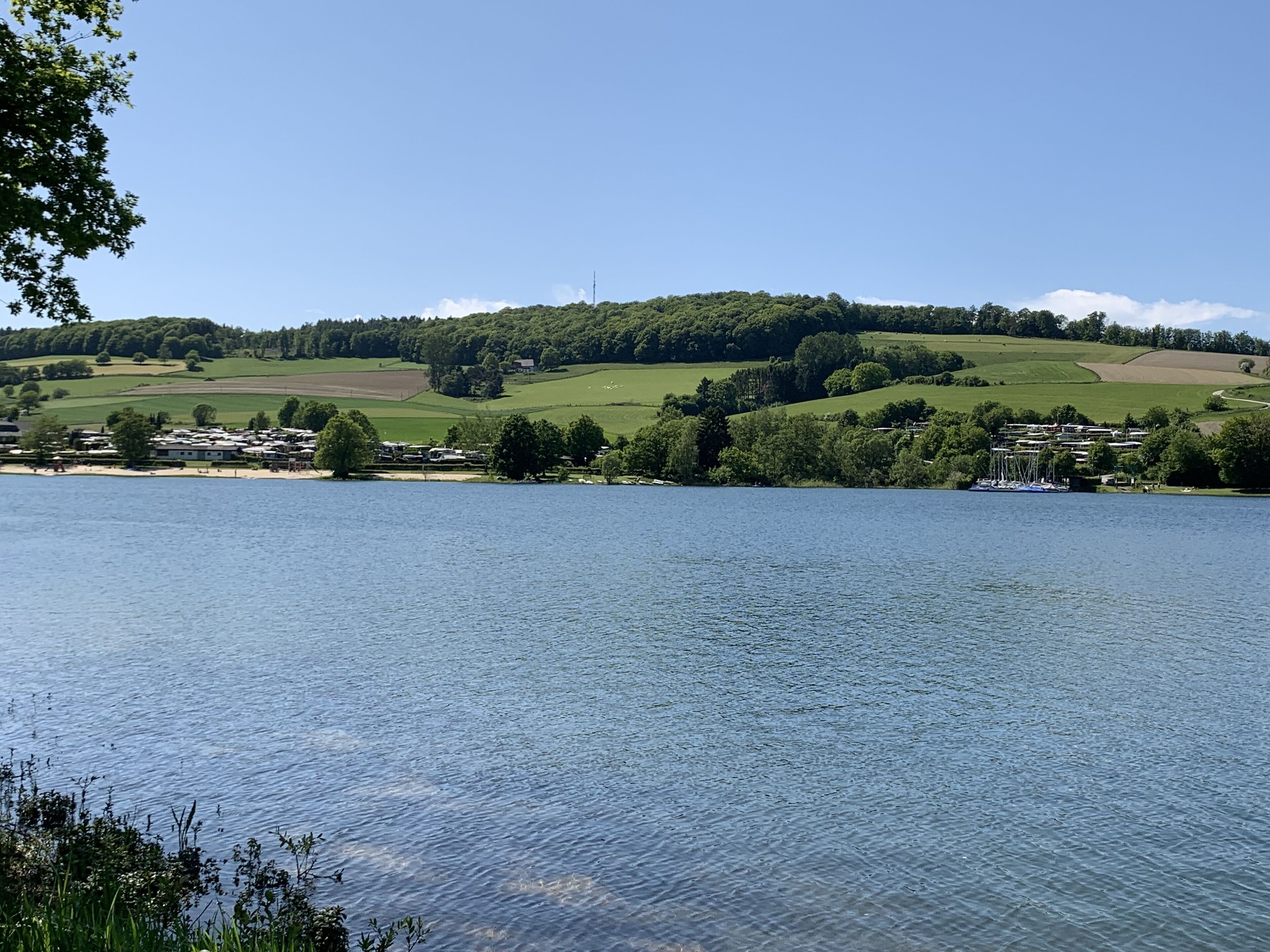 A lakeside view with a small settlement, including houses and boats, surrounded by green hills and clear skies.