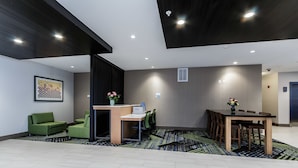 A modern hotel lobby with a reception desk, green seating, a dining area, and a patterned rug.