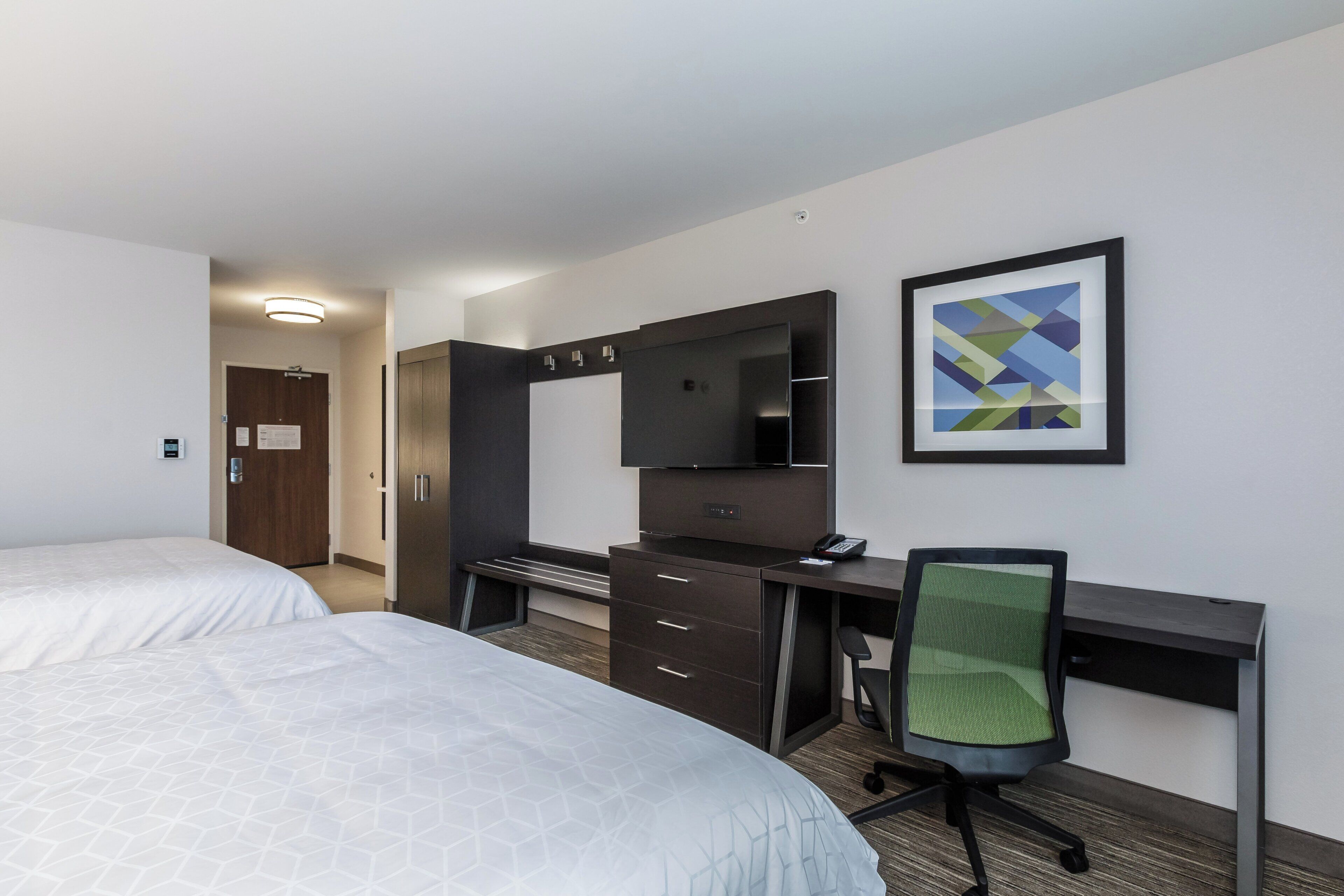 Habitación de hotel con dos camas, un escritorio, una silla, un espejo grande y un cuadro abstracto enmarcado.
