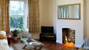 Una sala de estar con un sofá, una mesa de centro con libros y controles remotos, un televisor, una chimenea y un ventanal con cortinas.