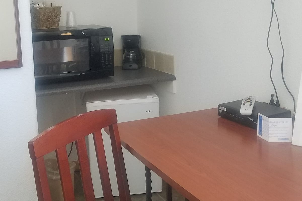 Uma pequena cozinha compacta com micro-ondas, máquina de café e torradeira. Há uma mesa de madeira com uma cadeira na frente.