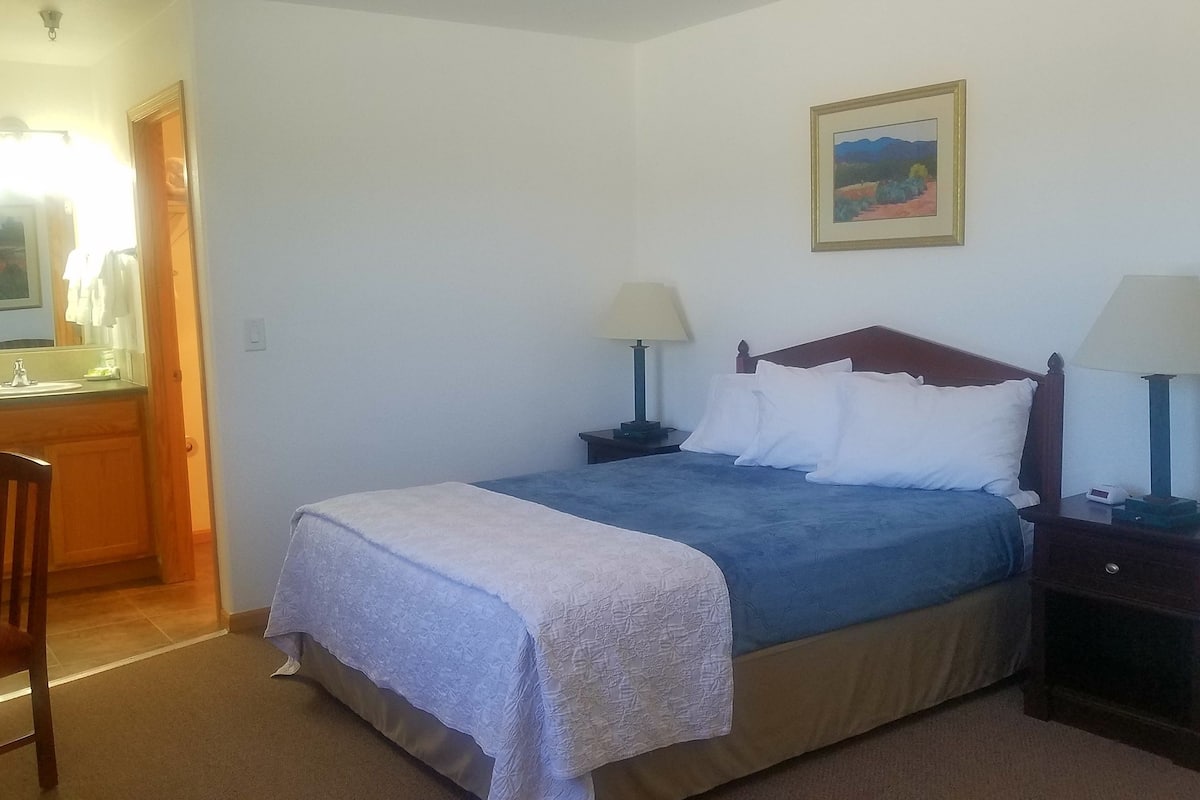 Um quarto com uma cama, duas mesas de cabeceira com luminárias, um quadro na parede e um banheiro visível por uma porta aberta.