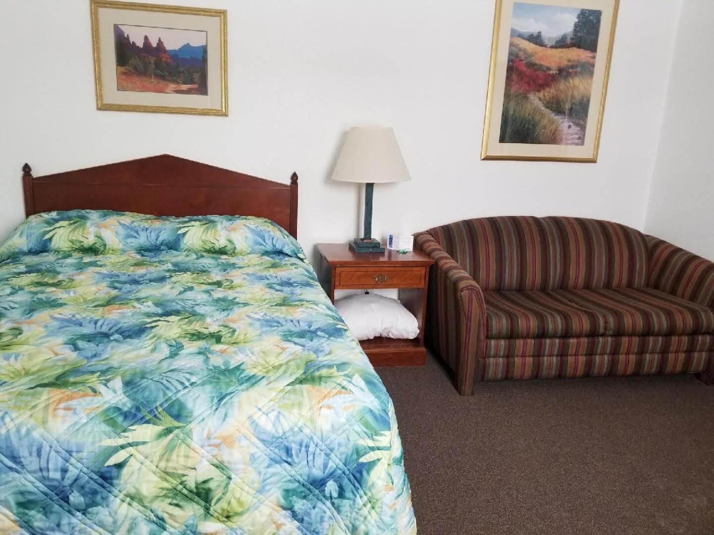 Un dormitorio con una cama, una mesita de noche, una lámpara, un sillón a rayas y dos cuadros en la pared.