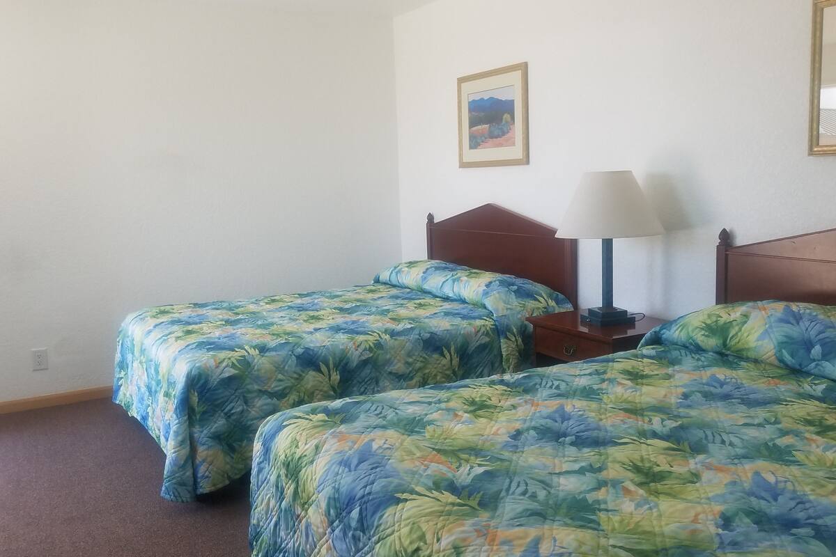 Quarto com duas camas de solteiro, cada uma com colcha floral, cabeceira de madeira, mesa de cabeceira com abajur e um quadro pendurado na parede.