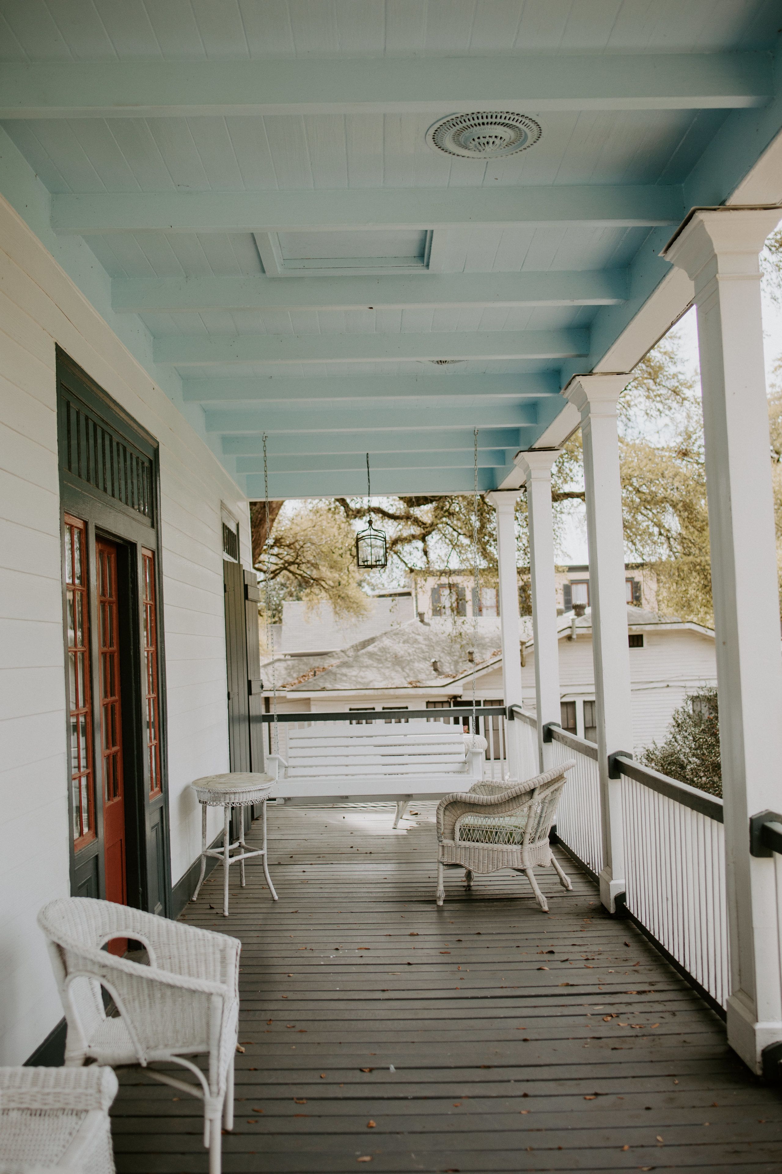 Une véranda couverte avec des chaises en osier peintes en blanc et une table, un plancher en bois et une vue sur une maison en arrière-plan.