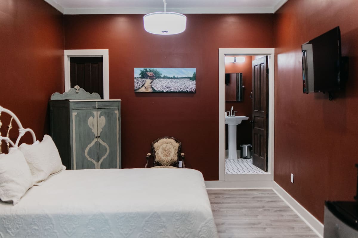 Une chambre à coucher avec un lit, une commode, une chaise, un tableau et une salle de bain visible à travers une porte ouverte.