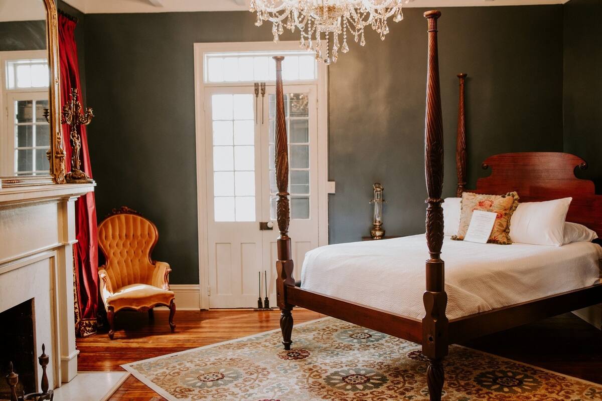 Un lit à baldaquin avec un couvre-lit blanc, une chaise en bois, une cheminée et un lustre.